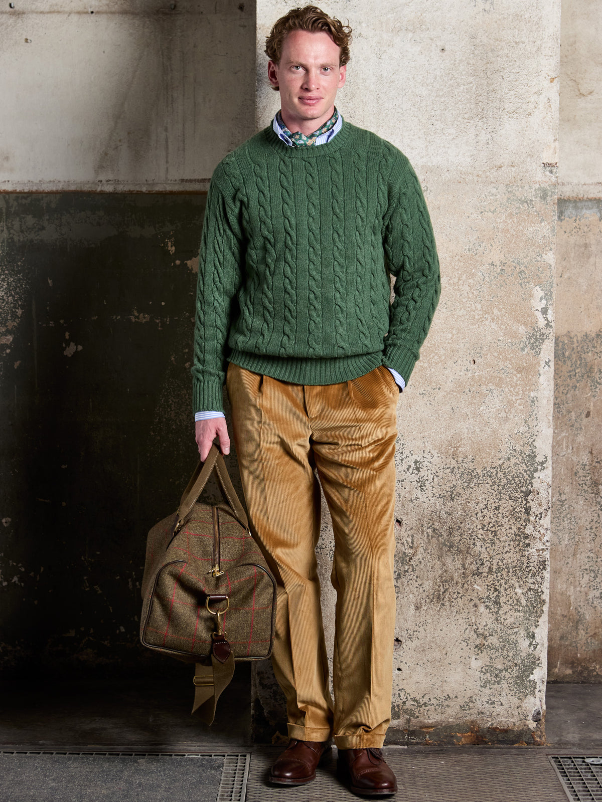Weekender aus Lovat Tweed in Olive in der Hand, kombiniert mit grünem Zopfpullover und Cordhose – John Crocket